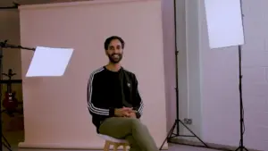 Smiling man in casual attire sitting on a stool against a soft background with studio lighting.