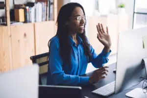 Woman with glasses waves while sitting at a desk, engaged in a video call with a computer in a stylish office.