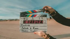 Film clapperboard held outdoors, marked with "Roadside" "Jakob & Ryan" and "Thomas Taught" under a blue sky.