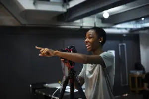 Smiling woman pointing while operating a camera on a tripod in a modern studio setting.