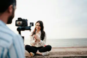 Woman sitting by the beach, smiling and recording a video on a camera with a scenic ocean background.