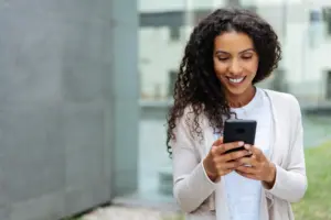 Smiling woman with curly hair using a smartphone outdoors, wearing a light sweater, enjoying a sunny day.