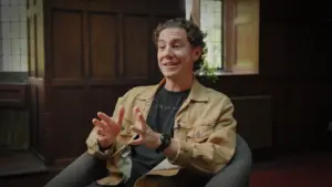 Person sitting in a modern armchair, gesturing while discussing ideas in a cozy, well-lit room with wooden paneling.