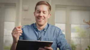 Person using a tablet in a bright workspace, wearing a denim shirt, focused on digital content creation.