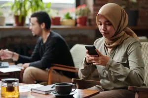 Muslim woman with a hijab using a smartphone in a cozy café, with a man working in the background.