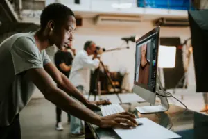 Young graphic designer working on a computer in a photography studio with others capturing images in the background.