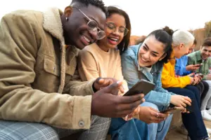 Group of friends enjoying time together, laughing and using smartphones outdoors in a lively social setting.