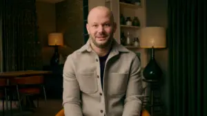 Bald man in a light gray jacket sits in a cozy, well-lit room with decorative lamps and shelves in the background.