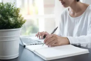 Woman writing in a notebook while working on a laptop, with a potted plant nearby for a fresh workplace vibe.