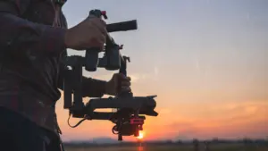 Person holding a camera stabilizer against a sunset backdrop, capturing the beauty of golden hour photography.