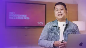 Man in denim jacket presenting on social media platforms in a modern office setting with a screen in the background.