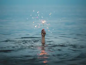 Person's hand emerging from water, holding a sparkling wand against a serene sky, creating a magical atmosphere at dusk.