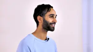 Smiling man in a light blue shirt with a microphone against a neutral background, showcasing a friendly demeanor.