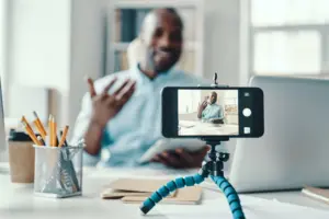 Man video conferencing on a smartphone, sitting at a desk with stationery and a laptop in a bright office space.