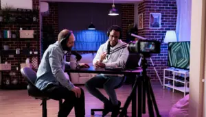 Two men recording a podcast in a modern studio with soft lighting, a camera, and a rustic brick wall backdrop.