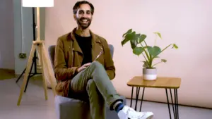 Smiling man sitting on a chair next to a plant and a table, dressed casually in a stylish indoor setting.