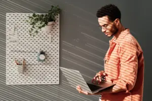 Young man using a laptop in a stylish workspace with wall planters and soft sunlight creating striped shadows.