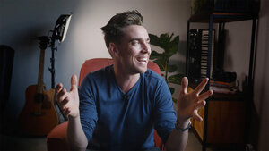 Smiling man gesturing enthusiastically while seated in a cozy, well-lit room with plants and musical instruments.