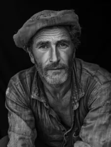 A somber portrait of a man in a worn-out shirt and a traditional hat, set against a dark background, evoking resilience.