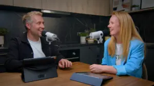 Two podcast hosts engaging in conversation at a modern kitchen table with microphones and digital devices.