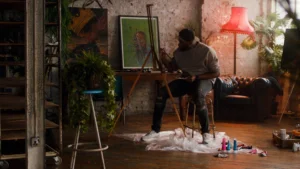 Artist sitting on a stool in a creative studio, painting on an easel with colorful art supplies nearby and plants in the background.