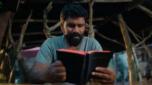 Man with a beard reading a book in a rustic setting, natural light illuminating his focused expression.