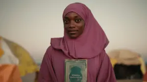 Young woman in a pink hijab holds a book, smiling against a soft background with tents, reflecting hope and resilience.