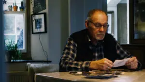 Elderly man examining old photographs at a table in a warmly lit, cozy living room with framed memories on the wall.