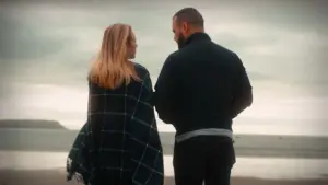 Couple walking on a beach, wrapped in a blanket, enjoying a serene moment by the ocean under a cloudy sky.