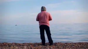 Person standing on a pebble beach, gazing at calm ocean waters under a clear blue sky.