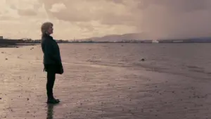 A woman stands by the shore under a cloudy sky, reflecting on the calm water and distant mountains.