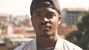 Young man in a baseball cap, looking confidently at the camera with a cityscape in the background.