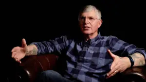 Man in a plaid shirt sitting on a leather couch, gesturing while speaking in a dimly lit setting.