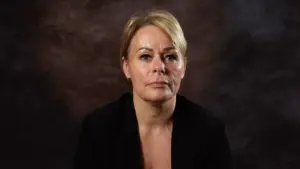 Portrait of a woman with short blonde hair, wearing a black blazer, against a dark textured background.