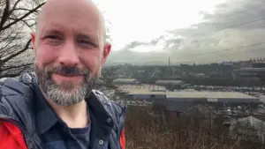 Smiling man in a jacket poses against a cloudy backdrop, overlooking a bustling industrial landscape.