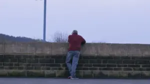 Man in a maroon shirt leaning against a stone wall, gazing at a distant landscape under a clear sky.