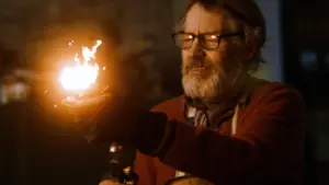 An elderly man with glasses holds a torch, showcasing flames in a dimly lit workshop, exuding a craftsman’s passion.