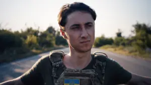 Soldier wearing tactical gear and a Ukrainian flag patch, standing on a rural road with a serious expression.