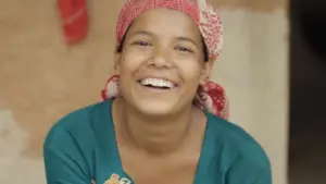 Smiling woman wearing a colorful headscarf, radiating joy and warmth in a vibrant rural setting.