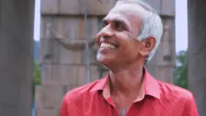 Elderly man in a red shirt standing between stone pillars, surrounded by greenery and historical architecture.