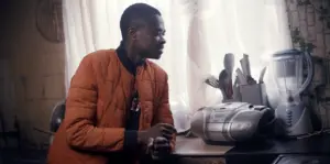 Young man in an orange jacket sitting near a boombox and blender, surrounded by a cozy, well-lit kitchen atmosphere.