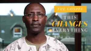 Man in traditional attire with glasses, background text reads "The Gospel: This Changes Everything."