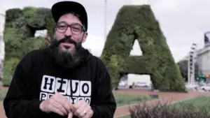 Man with a beard wearing a cap and hoodie stands in front of large greenery letters in an urban setting.