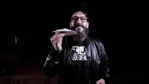 Man with a beard and glasses, wearing a leather jacket, holds a paper airplane against a dark background.