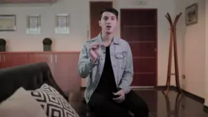 A young man in a denim jacket sits on a couch, gesturing while speaking in a stylish interior space.