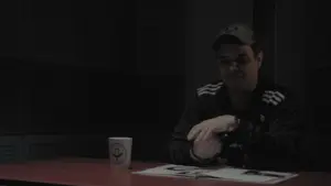 A man seated at a table in a dimly lit room, wearing a cap and casually examining documents with a coffee mug nearby.