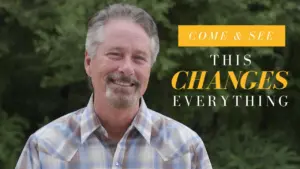 Smiling man in plaid shirt with greenery in background, promoting a life-changing experience.