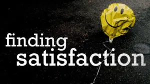 Yellow smiley balloon on dark surface with the text "finding satisfaction" creatively displayed beside it.