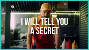 Man in an orange cap and red apron stands in a kitchen, ready to share a secret, with condiments in the background.