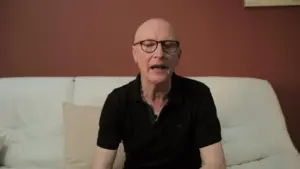 Bald man in glasses sitting on a couch, speaking against a warm-toned wall, casual attire and relaxed atmosphere.